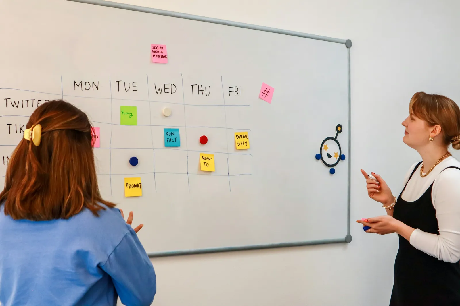 Two colleagues planning a week of social media posts on a whiteboard grid of days and channels.